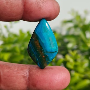 Könnte beinhalten: Ein polierter Edelstein in Diamantenform. Der Stein zeigt eine Mischung aus Türkis, Blaugrün und Brauntönen mit sichtbaren Bändern und Mustern. Der Edelstein wird zwischen zwei Fingern gehalten, vor einem verschwommenen grünen Hintergrund.