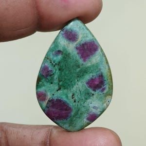 Puede incluir: Una gema en forma de lágrima con una base verde y parches irregulares de color rojo intenso. La superficie de la piedra tiene un aspecto moteado, con variaciones de color y textura. La gema está sujeta entre dos dedos.