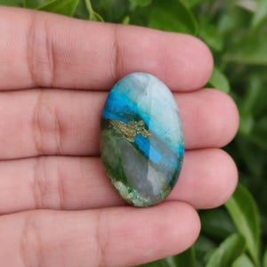 Op de afbeelding: Een ovale edelsteen met een gepolijst oppervlak. De steen heeft een mix van kleuren, waaronder blauw, groen en goud. De edelsteen wordt in een hand gehouden, met een vage groene achtergrond.