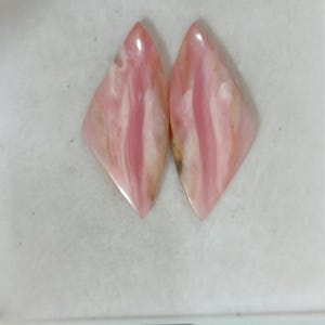May include: Two marquise-shaped pink opal cabochons featuring a smooth, polished surface with natural white and light pink veining patterns. These gemstones have a pointed top and bottom with a glossy finish, displaying organic colour variations.