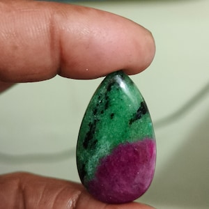 Può includere: Una pietra preziosa lucidata a forma di goccia con una base verde vibrante, inclusioni nere distinte e una sezione prominente di colore magenta intenso nella parte inferiore, caratterizzata da una superficie liscia e riflettente.