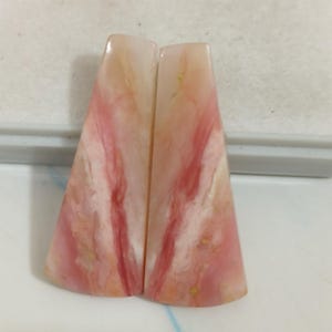 Può includere: Due cabochon trapezoidali in opale rosa lucido. Ogni pezzo presenta una superficie liscia e brillante con motivi vorticosi di crema pallido, pesca tenue e venature magenta vibranti che corrono verticalmente attraverso la struttura.