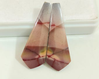 May include: Two polished gemstone pendants. Each pendant is a long, trapezoid shape with a faceted surface. The stones display a mix of colors, including shades of pink, red, yellow, and gray, with intricate veining patterns.