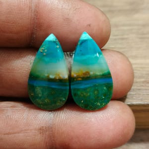 Op de afbeelding: Twee druppelvormige edelstenen. De stenen tonen lagen van blauwgroen, turquoise en groen, met vleugjes geel en bruin. De gepolijste oppervlakken reflecteren het licht en benadrukken de natuurlijke patronen.