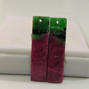 Pode incluir: Dois brincos retangulares de pedra preciosa. A parte superior é verde com manchas pretas, passando para um tom magenta profundo na parte inferior. Cada brinco tem um pequeno orifício na parte superior.