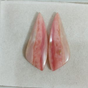May include: Two polished, teardrop-shaped pink opal gemstones featuring smooth, glossy surfaces with natural variations in light and dark pink hues, arranged side-by-side to display their elongated, pointed forms and distinct organic patterns.