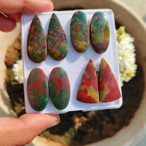 Pode incluir: Uma coleção de pedras preciosas polidas em várias formas, incluindo gotas, ovais e triângulos. As pedras exibem uma mistura de tons vermelhos, verdes e amarelos, com um padrão salpicado. As pedras preciosas são exibidas em um fundo branco.