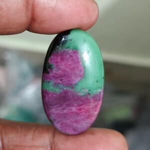 May include: An oval-shaped cabochon gemstone featuring a smooth, polished surface with a distinct colour pattern of vibrant magenta-pink patches contrasted against a bright green matrix, with a small area of deep black mineral inclusion at the top.
