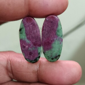 May include: Two oval-shaped cabochons featuring a natural blend of deep magenta-red ruby and light green zoisite. The stones display a smooth, polished surface with distinct colour zoning and small black mineral inclusions throughout the surface.