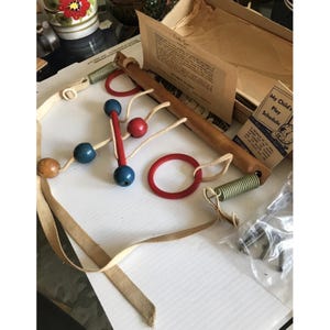 May include: Vintage wooden baby gym toy with red, blue, and natural wood elements. Includes a wooden bar, red rings, and a spring. The set also contains a box with printed instructions and a play schedule.