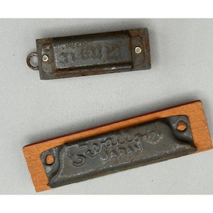 May include: Two vintage harmonicas. One is a small, rectangular, metal harmonica with the text "MIE JAPAN". The other is a similar harmonica with the text "Sawa JAPAN" on a wooden base.