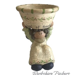 May include: A ceramic angel figurine holding a small bowl. The angel is dressed in a cream-colored dress with green accents and dark blue shoes. The bowl has a crackle finish with green and red floral details, and the words "Angel of Friendship".
