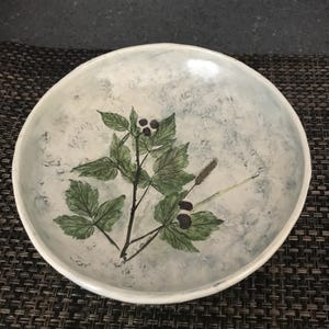 May include: A round, white ceramic bowl with a hand-painted botanical design. The design features green leaves, dark berries, and a light blue and gray background. The bowl has a slightly irregular edge.