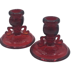 May include: Two ruby red glass candle holders. Each has a round base with a floral design, a central stem, and a cup-shaped top. The stems have decorative, curved arms. A classic home decor item.