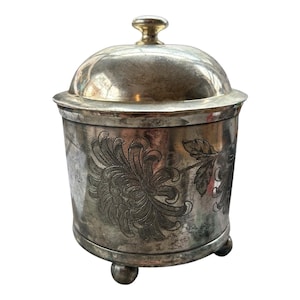 May include: A silver-toned container with a domed lid and a small, gold-colored knob. The container features an etched floral design and rests on three small ball feet. The container is likely used for storage.
