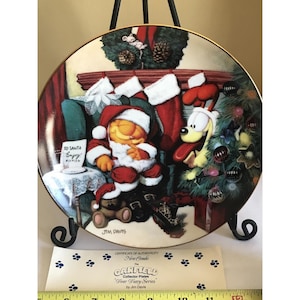 May include: A decorative collector's plate featuring Garfield in a Santa Claus outfit, sleeping near a Christmas tree. The plate has a gold rim and includes the text "To Santa Enjoy!" and "Fear Diary Series".