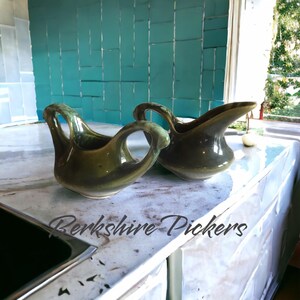 May include: Two olive green ceramic pitchers with unique handle designs. The pitchers have a glossy finish and a spout for pouring. The background features a teal tile backsplash and a countertop. The text "Berkshire Pickers" is visible.