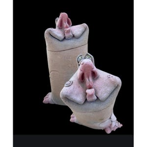 May include: Two whimsical ceramic sculptures of hippos. One is upright and cylindrical, the other is seated. Both have pink snouts, ears, and feet, with gray bodies and green eyes. The sculptures are on a black background.