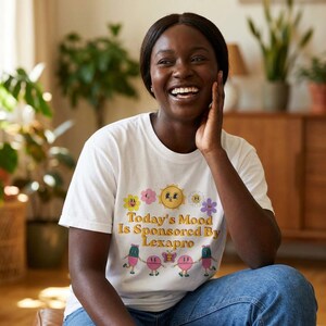 May include: A white t-shirt with the text "Today's Mood Is Sponsored By Lexapro" in a retro font, along with colorful flower and sun graphics. The person is wearing blue jeans.