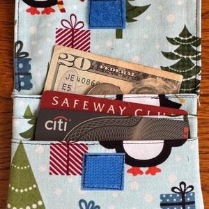 May include: A light blue fabric wallet with a Christmas theme. The wallet features a pattern of penguins, Christmas trees, and wrapped gifts. Inside, a twenty-dollar bill and two cards are visible. The wallet is secured with a blue square closure.