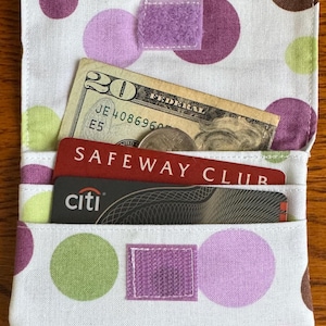 May include: A handmade fabric wallet with a white background and large polka dots in purple, green, and brown. The wallet is open, revealing a twenty-dollar bill, coins, and two credit cards. A purple velcro closure is visible.