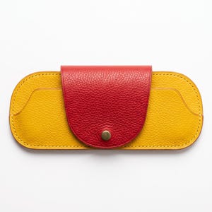 May include: A yellow leather glasses case with a textured surface and a rounded shape. It features a red leather flap closure with a centered brass-colored metal snap button. The edges are finished with visible stitching along the perimeter.