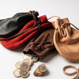 May include: A collection of four leather drawstring pouches in black, red, dark brown, and tan. The dark brown pouch is partially open, spilling several silver and gold-colored metal coins onto the surface. Each pouch features a matching leather cord.