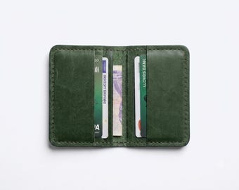 May include: A forest green leather card holder wallet featuring visible stitching along the edges. The open wallet contains multiple credit cards and a folded banknote tucked into the central compartment, displaying various bank logos and text.