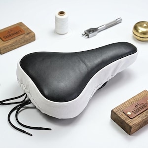 May include: A bicycle saddle featuring a black top and white sides with black lacing at the rear. The set includes a white spool of thread, a metal punch tool, a brass bell, and two wooden blocks with leather labels embossed with the text SAGENHAFT.