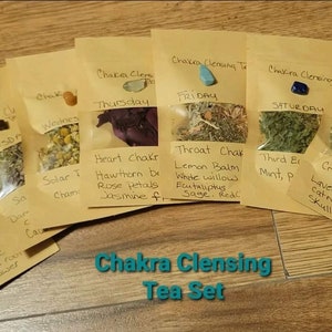 Op de afbeelding: Een set van zeven kleine bruine papieren zakjes met gedroogde kruiden en kristallen. Elk zakje is gelabeld met een dag van de week en de naam van een chakra. De zakjes zijn in een rij op een houten oppervlak gerangschikt. De tekst op de zakjes luidt: "Chakra Cleansing Tea Set".