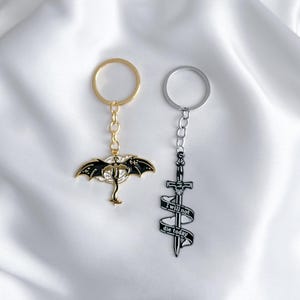 May include: Two keyrings with metal charms. One is gold-toned with a black and white dragon design. The other is silver-toned with a sword and a banner that reads "I will not die today".