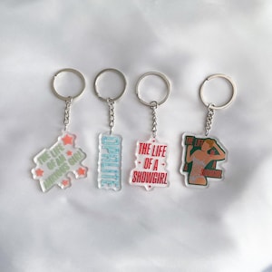 May include: Four keyrings with clear acrylic pendants. The pendants feature text and graphics, including "THE LIFE OF AN ANXIOUS GIRL" and "THE LIFE OF A SHOWGIRL". The keyrings have silver chains and rings. Graphics include stars and a showgirl illustration.