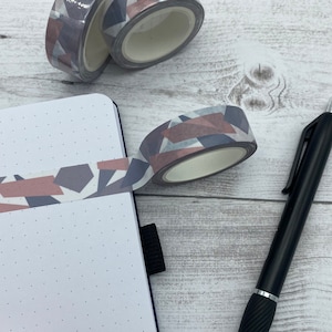 Könnte beinhalten: Zwei Rollen Washi-Tape mit einem geometrischen Muster in Grautönen und Rosa. Das Klebeband befindet sich auf einer weißen Holzoberfläche mit einem schwarzen Stift.