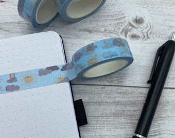 May include: A blue washi tape with a repeating pattern of clouds, rainbows, and suns. The tape is rolled up and placed on a white surface next to a black pen and a notebook.