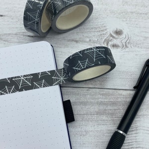 Op de afbeelding: Een zwart-wit gepatroneerd washi tape met een spinnenwebontwerp. De tape is opgerold en geplaatst op een wit oppervlak naast een notitieboek.