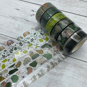 May include: A set of six washi tapes with various green and brown floral and geometric patterns. The tapes are rolled up and stacked on top of each other. The tapes are on a white wooden surface.