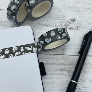 May include: A roll of black and white washi tape with a pattern of white ghosts on a black background. The tape is partially unrolled and is being used to decorate a notebook.
