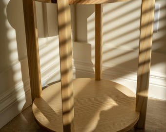 May include: A light wood end table with a round top and a lower shelf. The table has a simple, modern design and is made of natural wood.
