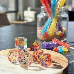 May include: A set of seven multi-faceted polyhedral dice made of clear resin with embedded colorful numbers and shapes. The dice set includes a d20, d12, d10, d8, d6, and d4, arranged in a cluster. The numbers are printed in various bright colors.