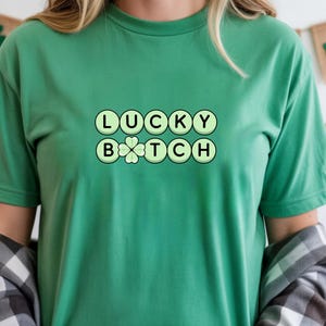 Può includere: Una maglietta verde con la scritta "LUCKY BITCH" in un carattere giocoso e arrotondato. La parola "LUCKY" è sopra "BITCH", con un quadrifoglio che sostituisce la "O" in "BITCH". La maglietta è di colore verde unito.