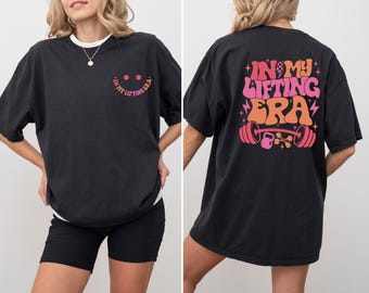 May include: Black oversized t-shirt with the text "IN MY LIFTING ERA" in pink and orange on the back, and a smiley face with the same text on the front. The shirt features a crew neck and short sleeves.