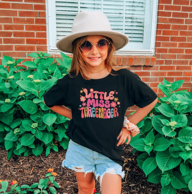 May include: A black t-shirt with the text "Little Miss Three-nager" in colorful letters and flowers. The shirt is being worn by a child wearing a brown hat and sunglasses.