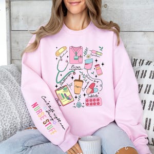 May include: A light pink sweatshirt featuring a colorful graphic of medical tools and phrases. The design includes a stethoscope, nurse uniform, coffee cup, and the words "NURSE STUFF". The sleeve has the text "Can't talk right now Doing".