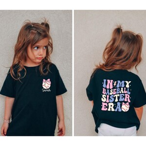 Może przedstawiać: Czarny T-shirt z r&oacute;żowym nadrukiem baseballu i imieniem "Sarah" z przodu. Z tyłu T-shirt ma napis "In My Baseball Sister Era" w kolorach tęczy z r&oacute;żowym nadrukiem baseballu.