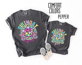 May include: Two matching charcoal gray t-shirts with a colorful smiley face design. The larger shirt says "Birthday Squad" and the smaller shirt says "Birthday Girl". The shirts have rolled sleeves and are paired with black and white checkered slip-on shoes.