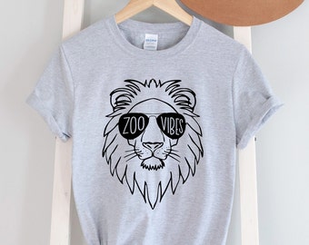 May include: A grey t-shirt featuring a black line drawing of a lion's head wearing sunglasses. The sunglasses have the text "ZOO VIBES" written on them.