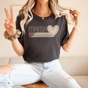 May include: A dark grey t-shirt with a retro-style graphic that says "Mama" with a pink, green, and gold heart. The text below the heart says "Hardworking, Protective, Selfless, Love".