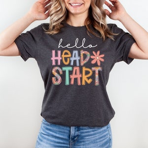 May include: A dark gray t-shirt with the text "hello head start" in colorful letters with floral designs. The letters are outlined in white.
