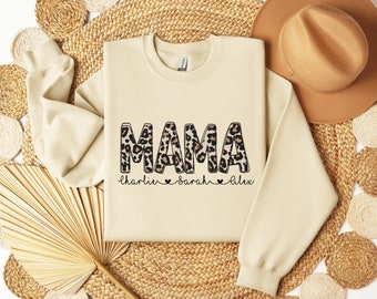 May include: Cream-colored sweatshirt with the word "MAMA" in leopard print letters. Below the word "MAMA" are the names "charlie", "sarah", and "alex". The sweatshirt is made of a soft material and has a crew neck.