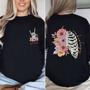 Pode incluir: Camiseta preta de manga comprida com um design floral e uma caixa tor&aacute;cica de esqueleto nas costas. O texto "radiologia" est&aacute; escrito verticalmente do lado direito da caixa tor&aacute;cica. A frente da camiseta tem uma m&atilde;o de esqueleto fazendo o sinal de rock and roll com flores ao redor. O texto "Sarah" est&aacute; escrito abaixo da m&atilde;o.